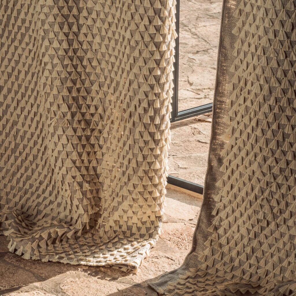 Close-up van Elitis Baleares Finca LZ 897 04 gordijnstof met een reliëfstructuur van beige driehoekige patronen in zonlicht.