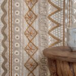 Detail van de Elitis Baleares Menorca LZ 899 03 stof met decoratieve borduursels in aardetinten naast een rustieke houten tafel en keramieken beker.