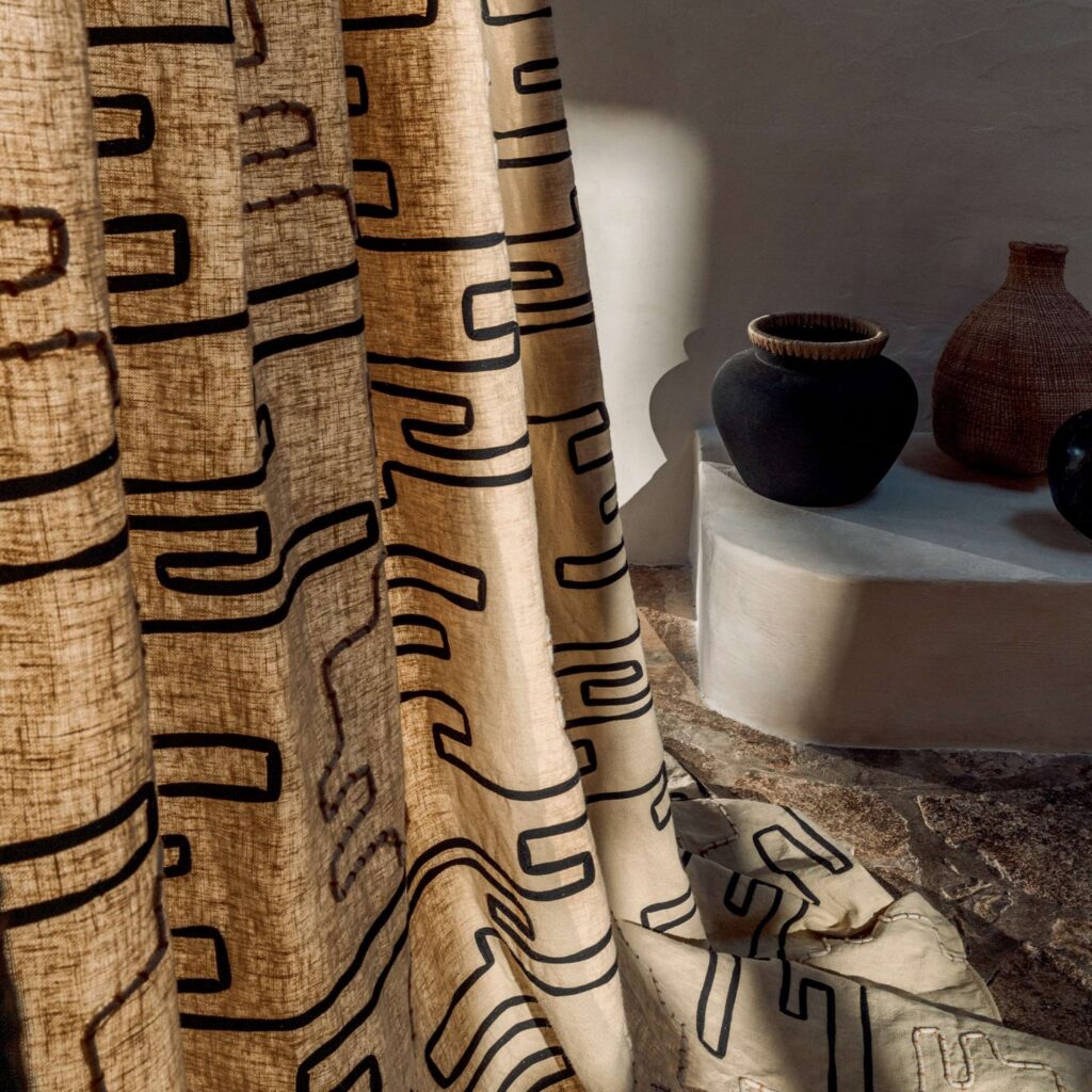 Beige Elitis Baleares Pacha gordijn met zwart abstract patroon in een sfeervol interieur met aardewerken vazen.