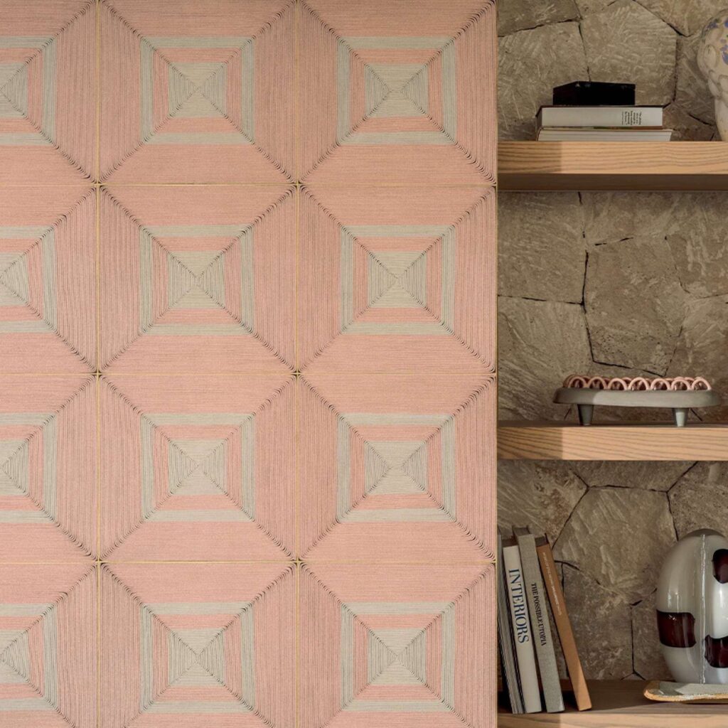 Decoratief wandpaneel uit de Elitis Cordes Sensibles-collectie met geometrisch patroon in roze- en natuurtinten naast een houten plank met boeken en woonaccessoires.
