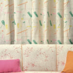 Crèmekleurig Elitis Excentric Bowie gordijn met abstract gekleurde patronen achter een licht gespikkelde bank met roze en oranje kussens.