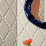 Reliefbehang Elitis Forms Wall Mone Lin RM 1057 01 met ruitpatroon, een blauwe organische spiegelrand en een oranje bloem op de voorgrond.