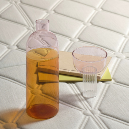 Roze glazen karaf en glas op een boek, geplaatst op een crèmekleurige Elitis Forms wandbekleding met reliëfpatroon.