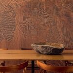 Houten tafel met houten schaal ervoor, tegen een muur met het reliëfpatroon van het Elitis Bois Sculpté Nuit Cinétique panorama.
