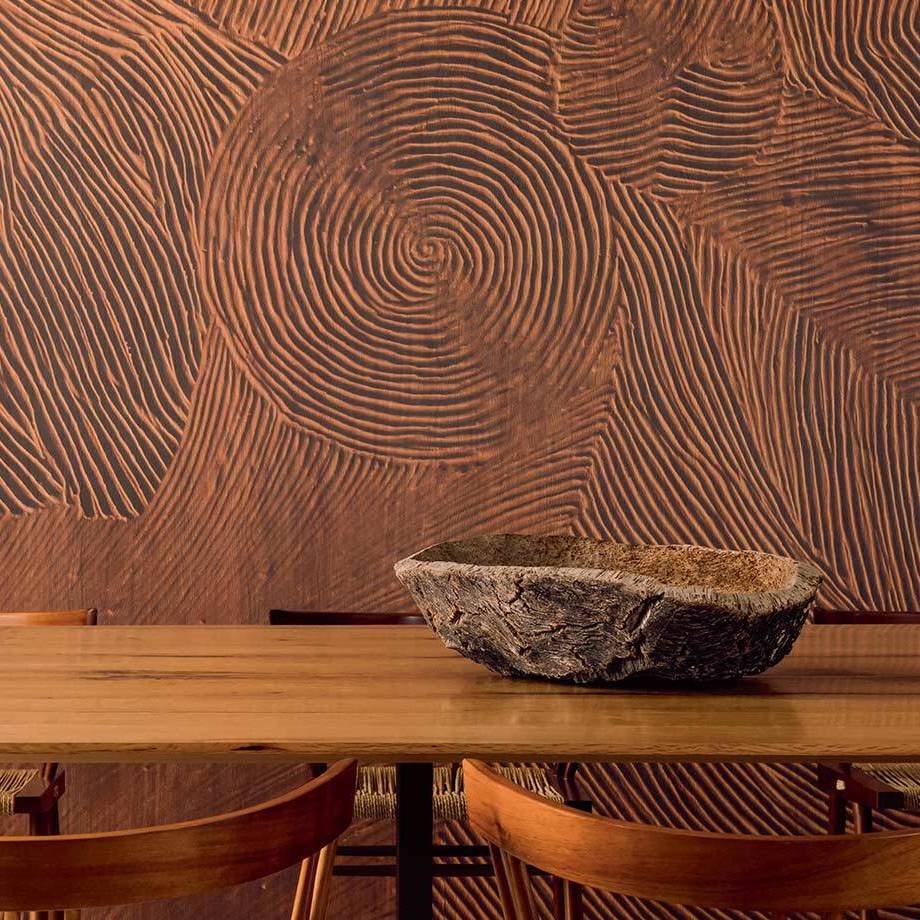 Houten tafel met houten schaal ervoor, tegen een muur met het reliëfpatroon van het Elitis Bois Sculpté Nuit Cinétique panorama.