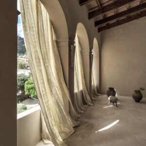 Lichte, golvende Elitis-stof Ephemere Univers LI 895 02 hangt voor hoge boogramen in een mediterraan interieur met natuurstenen vloer en houten plafonds.