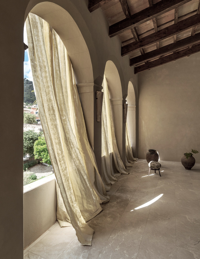 Lichte, golvende Elitis-stof Ephemere Univers LI 895 02 hangt voor hoge boogramen in een mediterraan interieur met natuurstenen vloer en houten plafonds.
