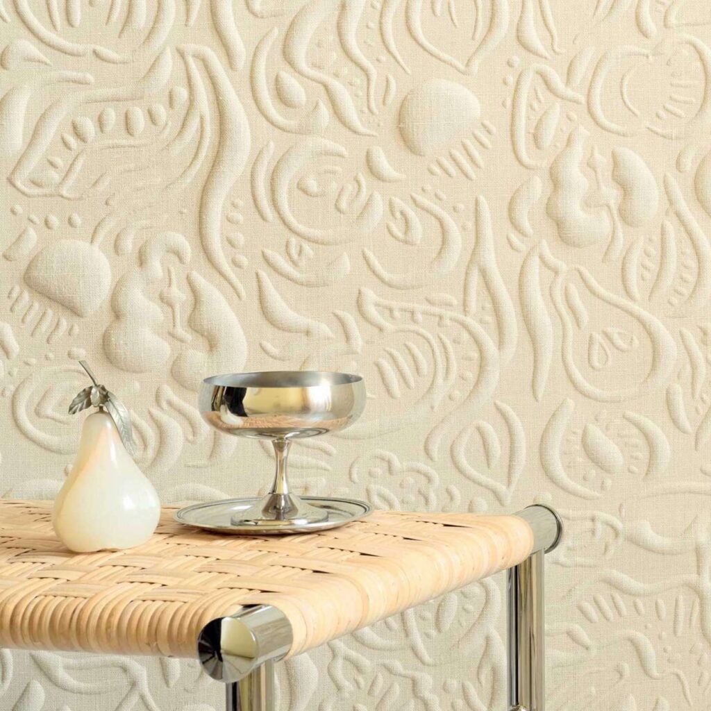 Decoratieve wand met het reliëfpatroon 'Le Jardin d’Aloès' in beige linnenlook, met een metalen tafeltje waarop een zilverkleurige schaal en een glanzend peervormig object staan.