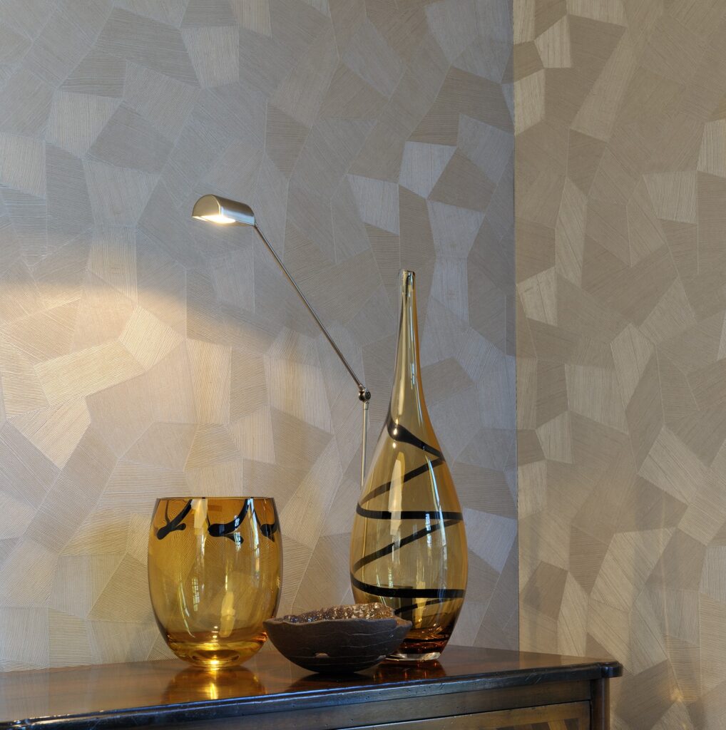 Glazen decoratieobjecten en een moderne leeslamp staan op een kast tegen een beige Arte Monsoon Facet 75301 wandbekleding met geometrisch patroon.