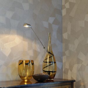 Glazen decoratieobjecten en een moderne leeslamp staan op een kast tegen een beige Arte Monsoon Facet 75301 wandbekleding met geometrisch patroon.