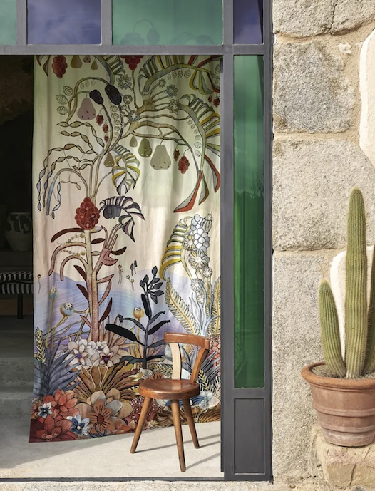 Gordijn uit de Christia Lacroix stoffen collectie 2025 met kleurrijk botanisch patroon hangt in een stenen deuropening naast een houten stoel en een cactus.