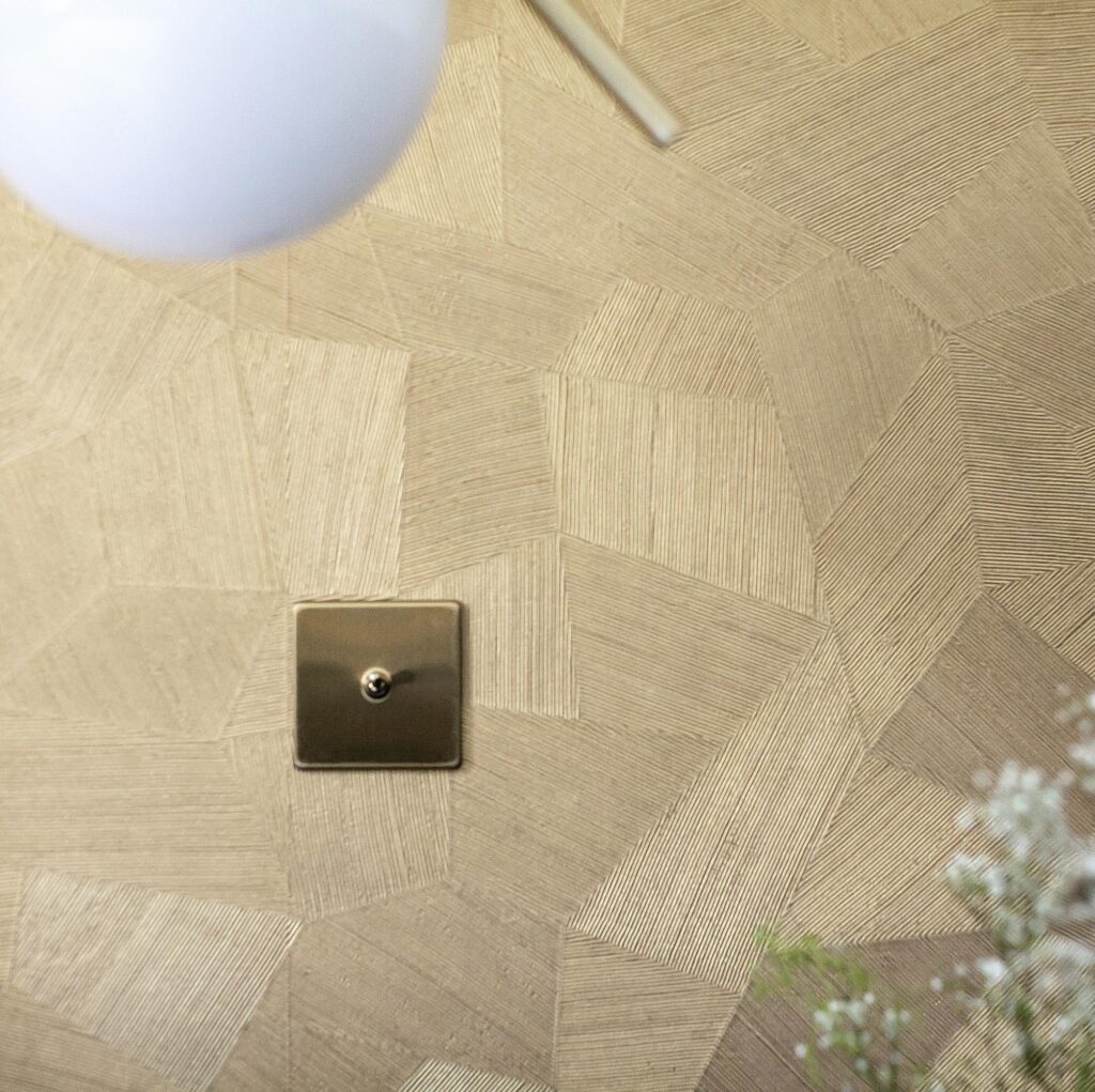 Detail van het Arte Facet 26543 behang met geometrisch patroon in beige en goudtinten, een lichtschakelaar en een deel van een lamp zichtbaar.