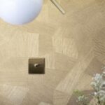 Detail van het Arte Facet 26543 behang met geometrisch patroon in beige en goudtinten, een lichtschakelaar en een deel van een lamp zichtbaar.