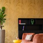 Sfeervol interieur met Arte Musa gold leaf behang, een fluwelen oranje fauteuil, een zwarte kast en groene kamerplant.