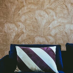 Donkerblauwe bank met een sierkussen voor een gouden Arte Sabal-structuurbehang met bladmotief.