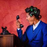 Man in blauwe blazer met haarrollers die boos in een ouderwetse telefoon schreeuwt, tegen een rode Arte Sabal behangachtergrond.