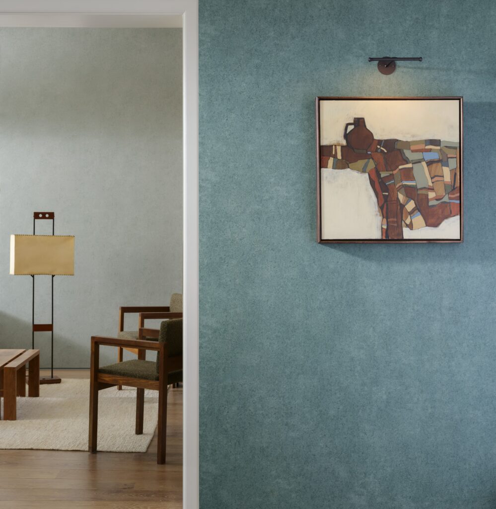 Moderne interieursetting met Arte Essentials Kaolin Calcaire Stucco behang in blauwgroene tint, een abstract schilderij en houten meubels.