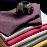 Gestapelde Elitis Kaila stoffen in diverse kleuren met een rustiek keramieken kommetje erbovenop.