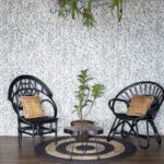 Twee zwarte rotanstoelen met gevlochten kussens staan op een rond vloerkleed voor een wand met Arte Moooi mo2041 behang, naast een kleine plant en organisch gevormd tafeltje.