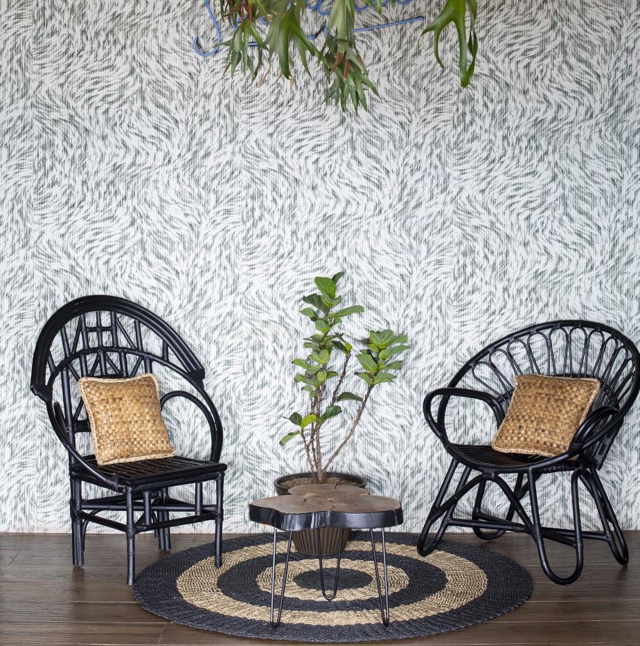 Twee zwarte rotanstoelen met gevlochten kussens staan op een rond vloerkleed voor een wand met Arte Moooi mo2041 behang, naast een kleine plant en organisch gevormd tafeltje.